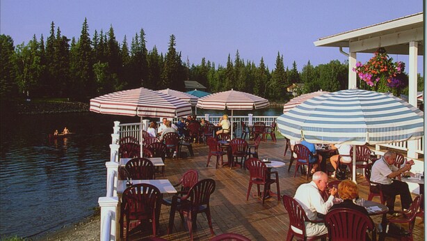 An outdoor dining area with tables and chairs, striped umbrellas, and a view of a lake. - Rivers Edge Resort (Fairbanks)