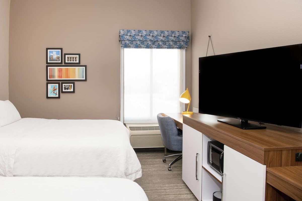 Une chambre d’hôtel avec deux lits, une télévision, un bureau avec une lampe et une fenêtre avec des stores.