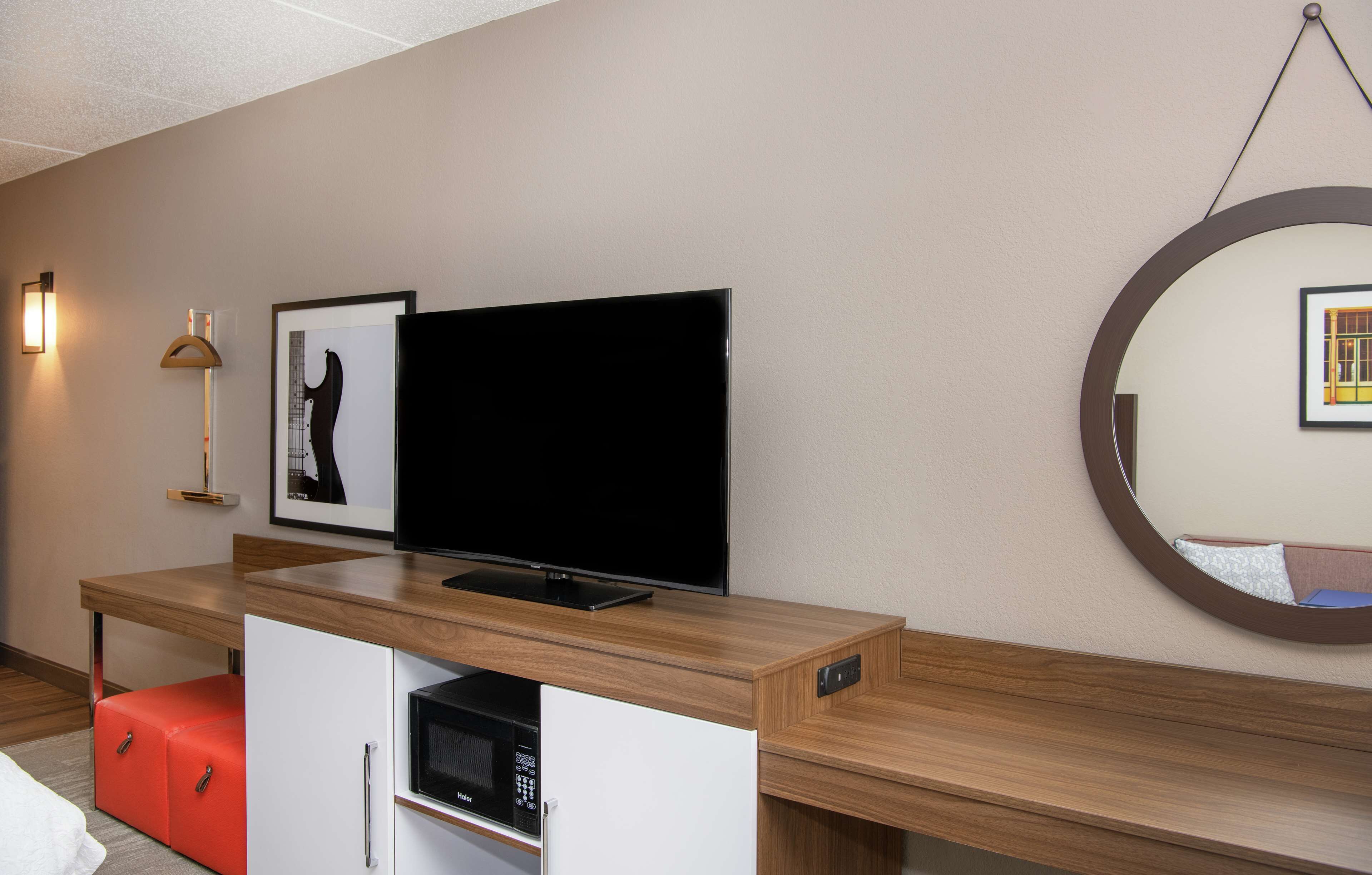 A modern hotel room with a wooden desk, a flat-screen TV, a microwave, a red ottoman, a framed picture, a wall-mounted lamp, and a round mirror.