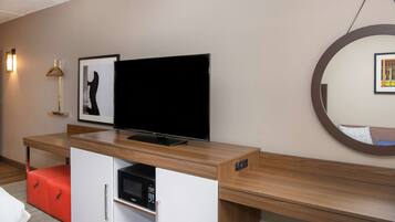 A modern hotel room with a wooden desk, a flat-screen TV, a microwave, a red ottoman, a framed picture, a wall-mounted lamp, and a round mirror.