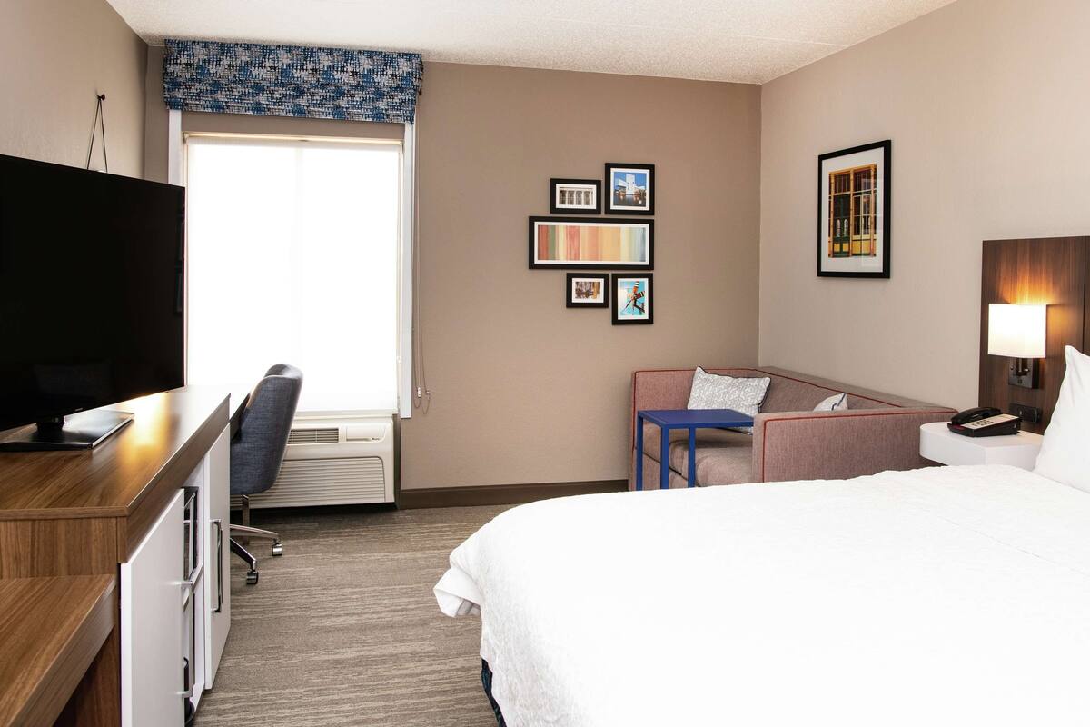 Une chambre d’hôtel comprenant un lit, un bureau avec une télévision, une chaise, une petite table et un mur orné de tableaux.