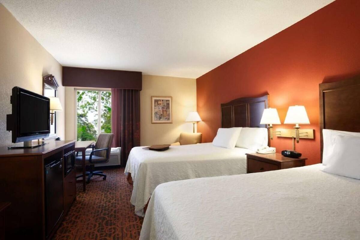 Une chambre d’hôtel avec deux lits, un bureau avec une télévision, une chaise, une fenêtre avec des rideaux et un tableau au mur.