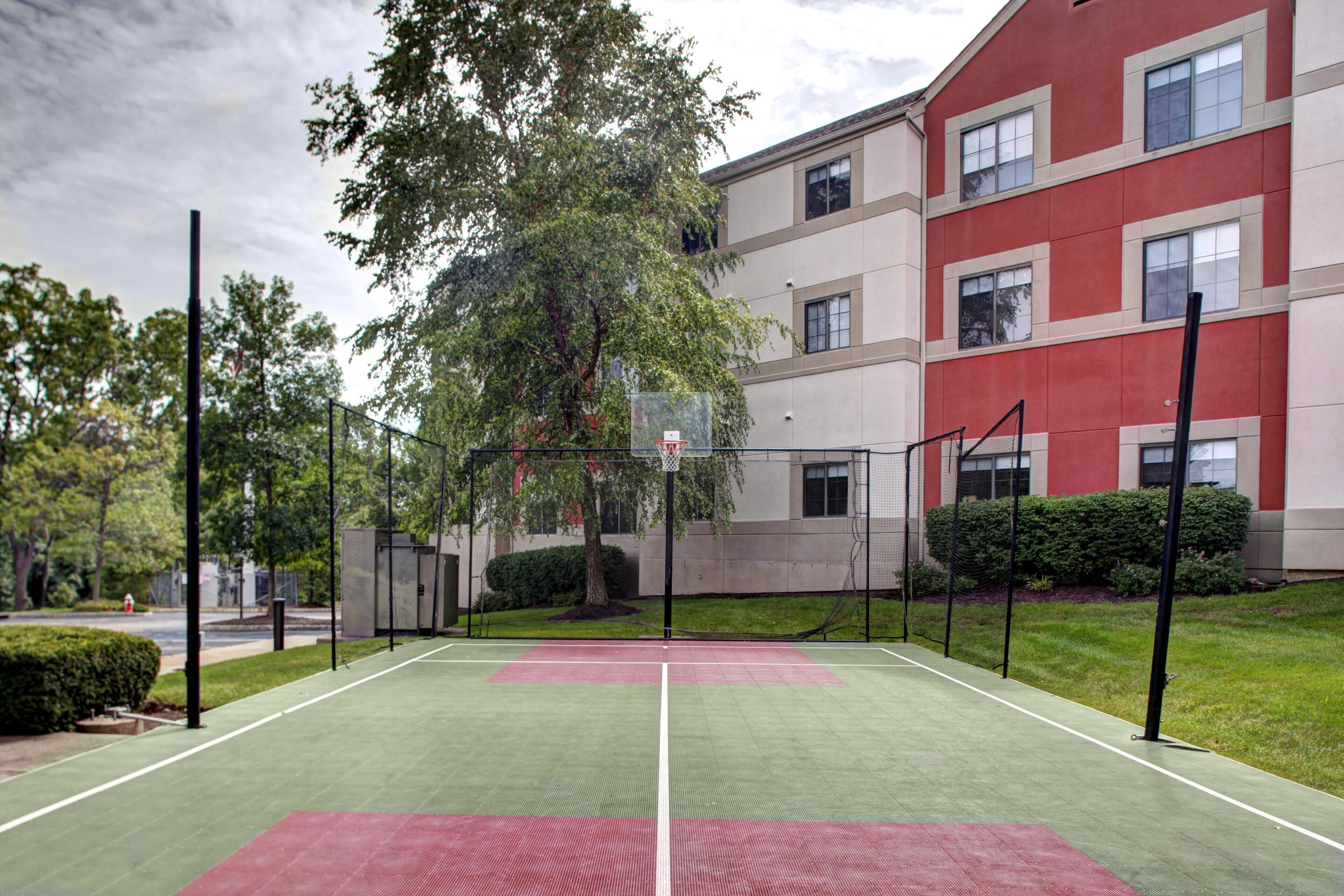 An outdoor basketball court with a net, surrounded by trees and a building with multiple windows.