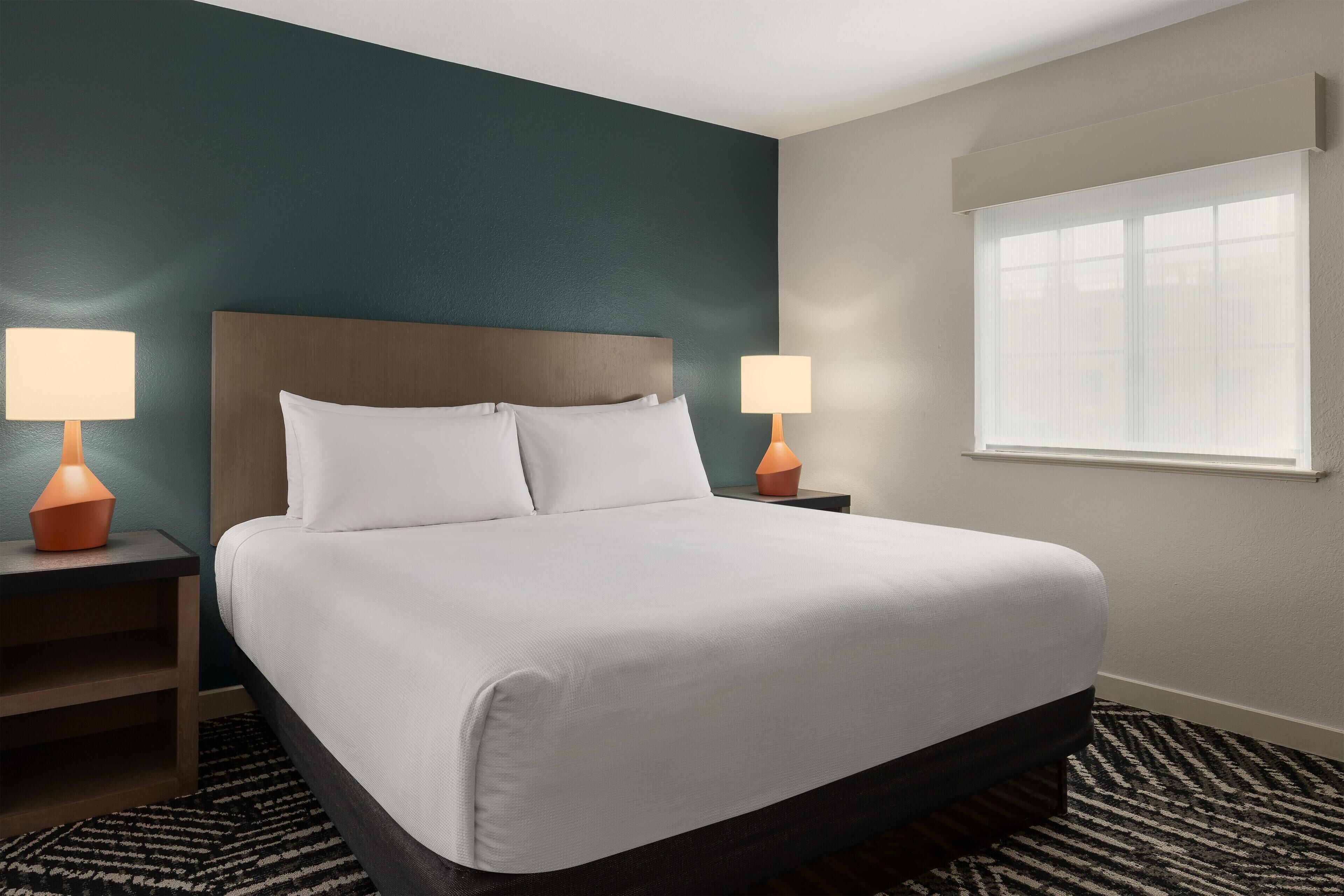 A neatly made bed with white linens, two bedside lamps, a window with blinds, and a dark wooden headboard.