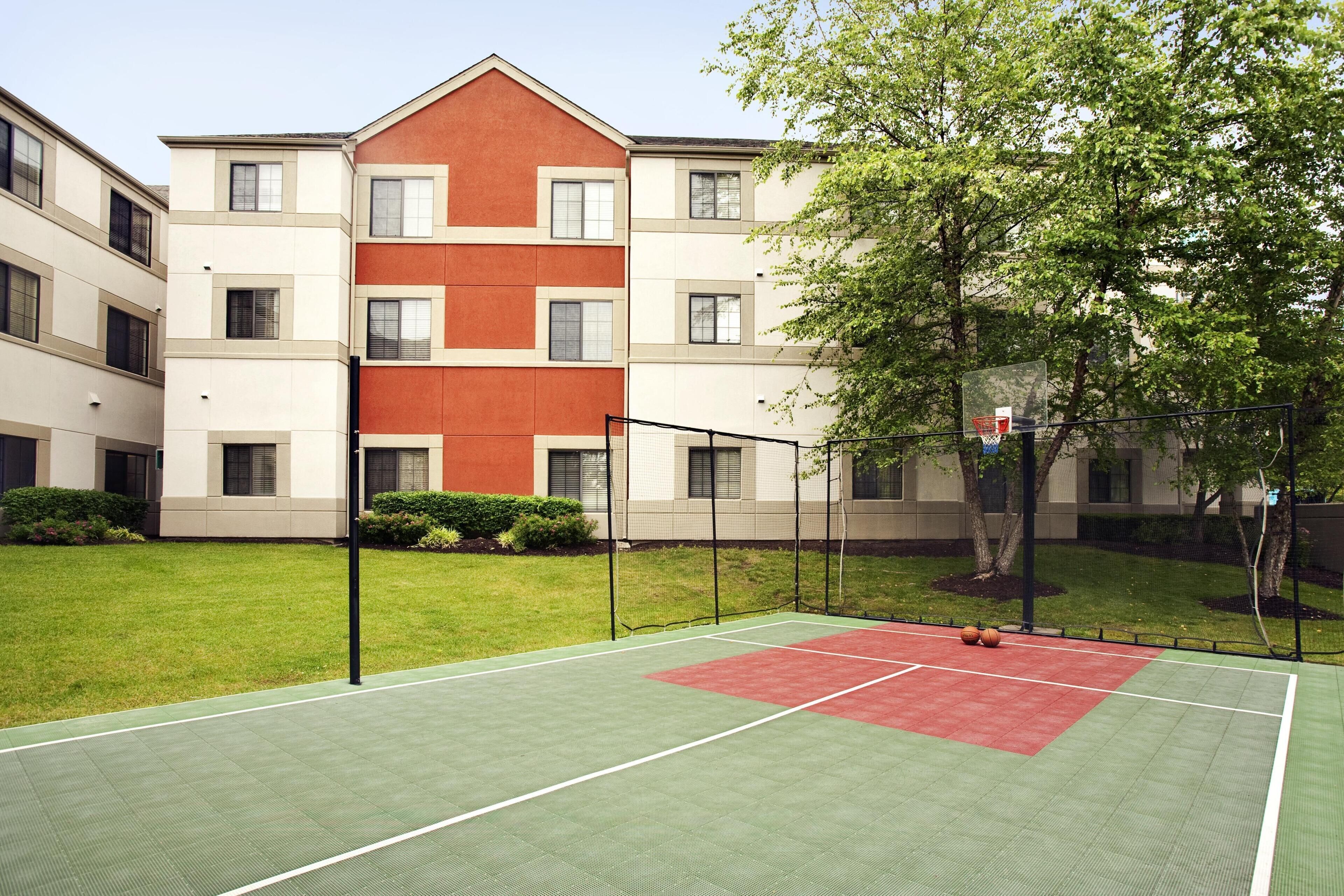 Un terrain de basket en plein air, équipé d’un panier, entouré d’une pelouse verdoyante et d’un bâtiment doté de nombreuses fenêtres.