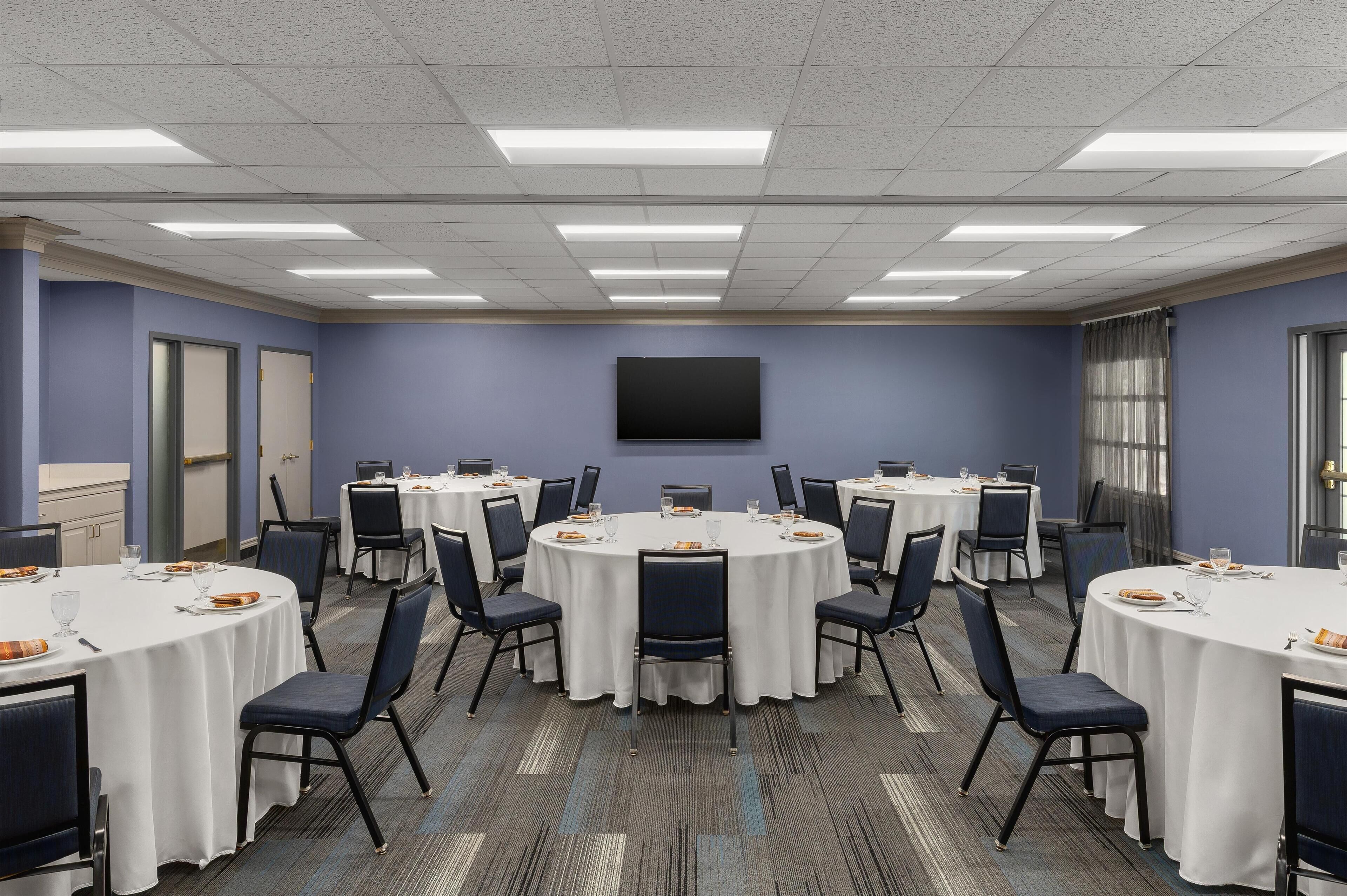A conference room with round tables, chairs, a flat-screen TV, and a window with curtains.