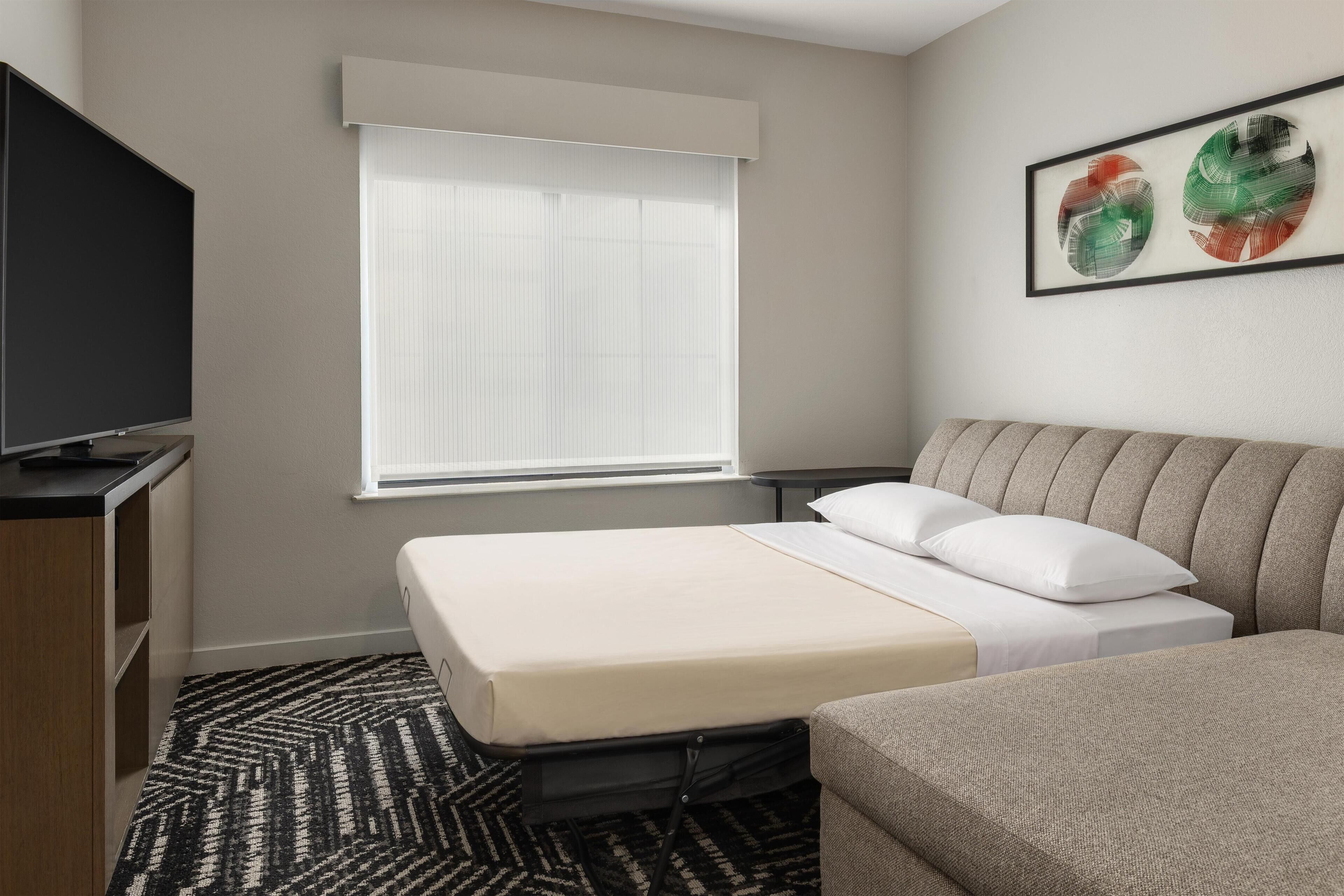 Een hotelkamer met een bed, een televisie, een raam met jaloezieën, een tafeltje en een schilderij aan de muur.