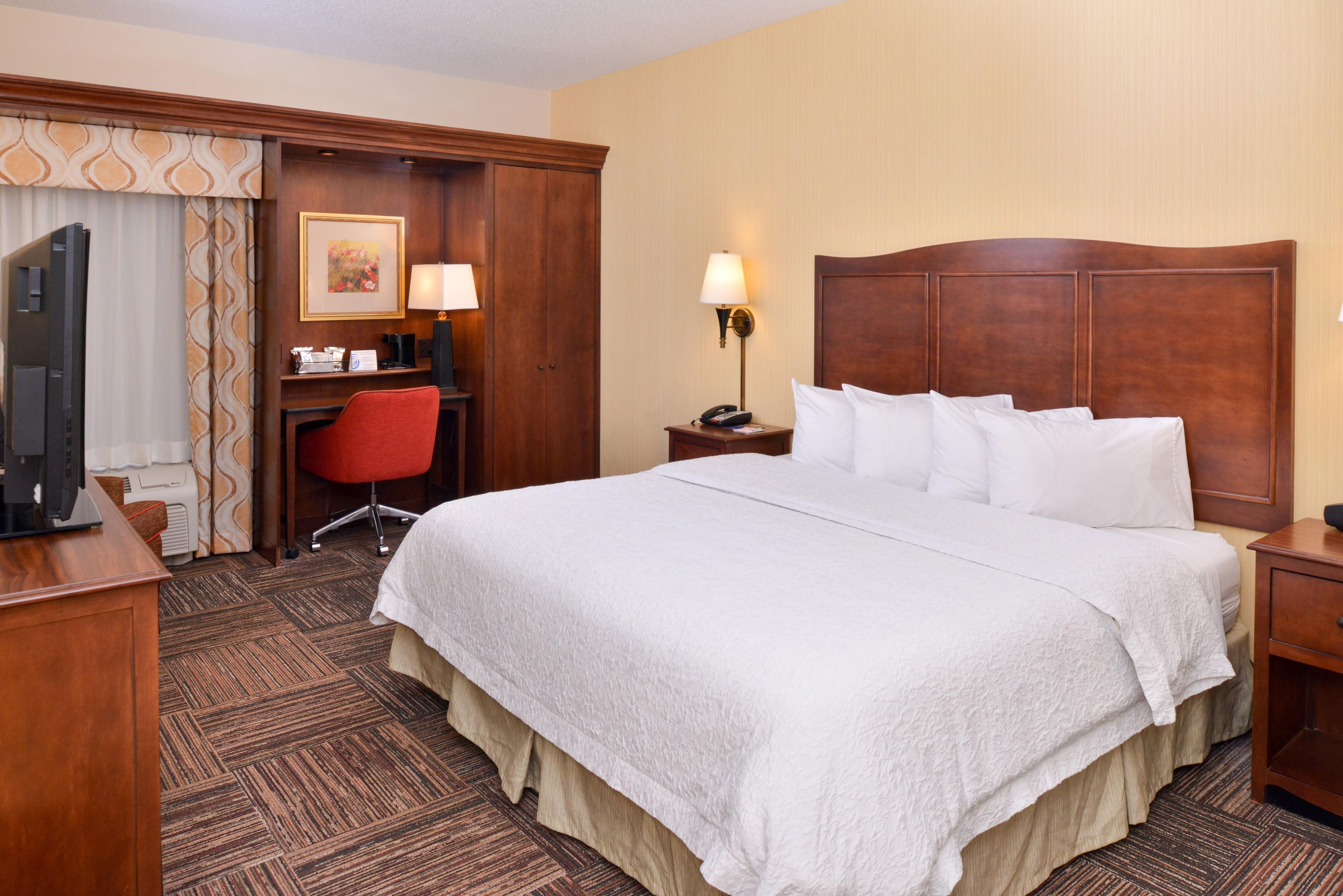 A hotel room with a large bed, a desk with a chair, a television, a desk lamp, and a nightstand with a lamp.