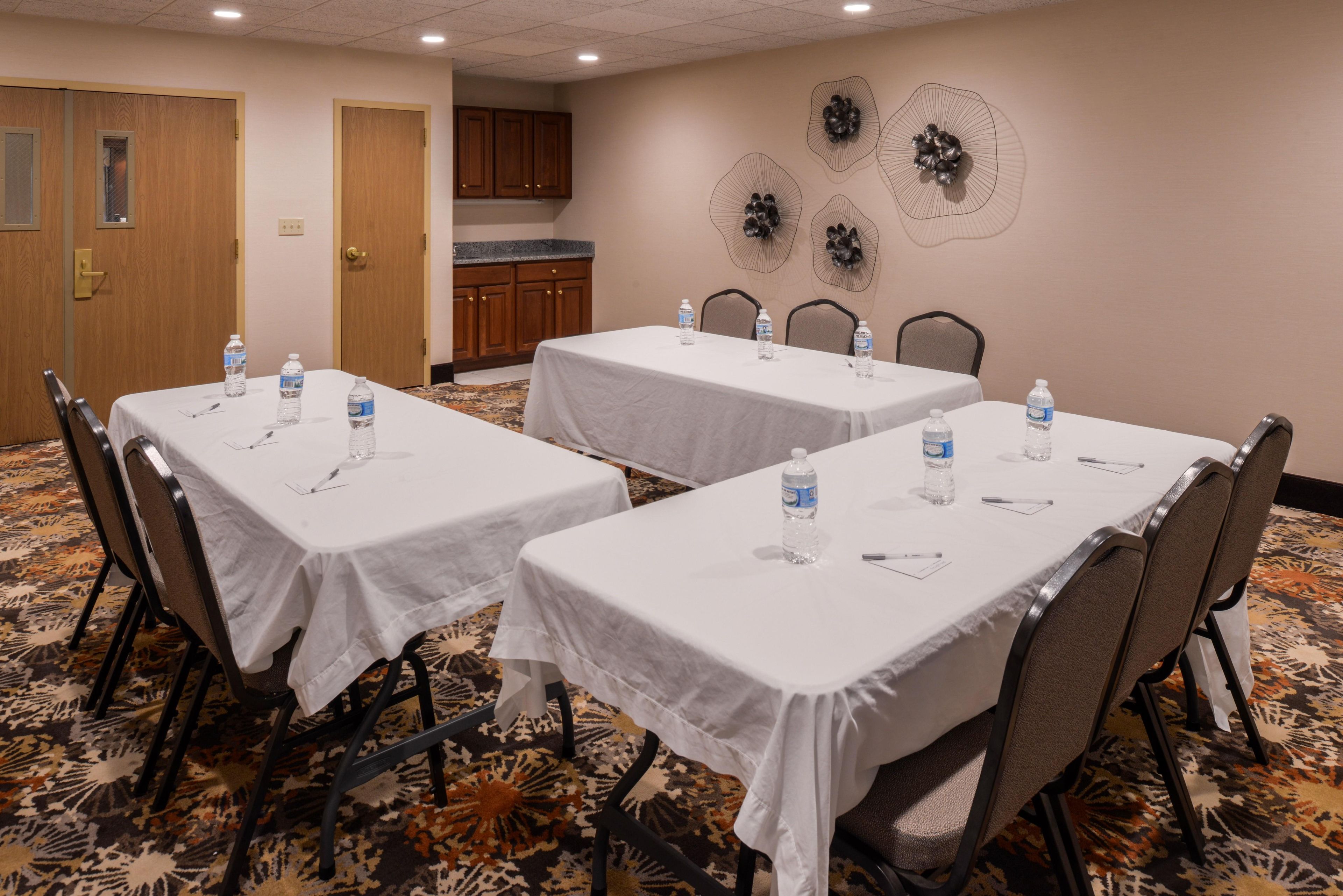Une salle de conférence avec trois tables, chacune recouverte d’une nappe blanche, des chaises et des bouteilles d’eau.