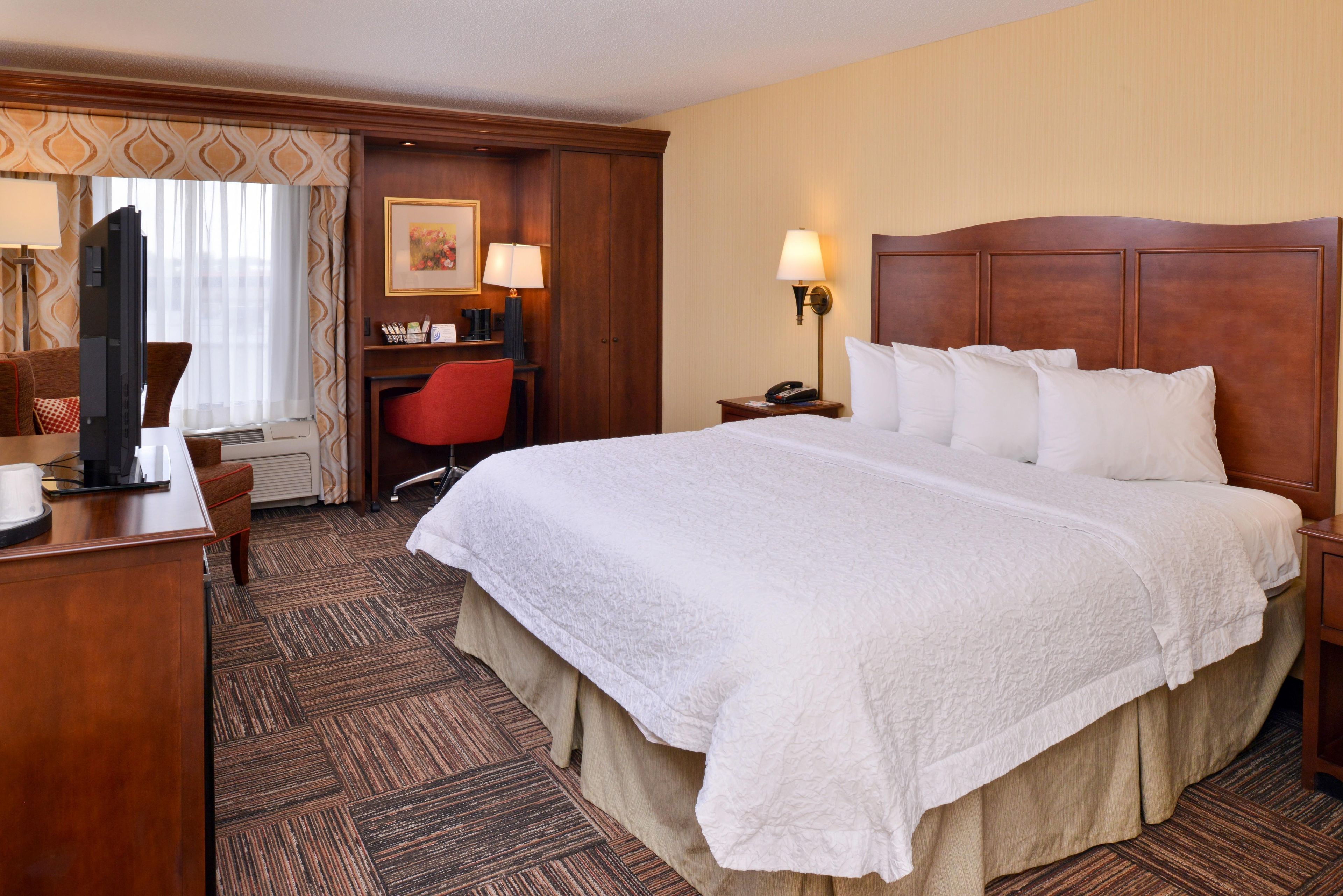 Quarto de hotel com uma cama grande, uma escrivaninha, uma cadeira, uma televisão e um abajur.