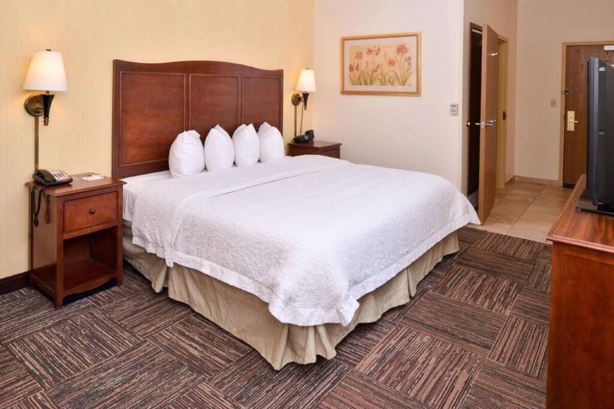 A hotel room with a large bed, two bedside tables with lamps, a wooden headboard, a painting on the wall, and a door to the right.