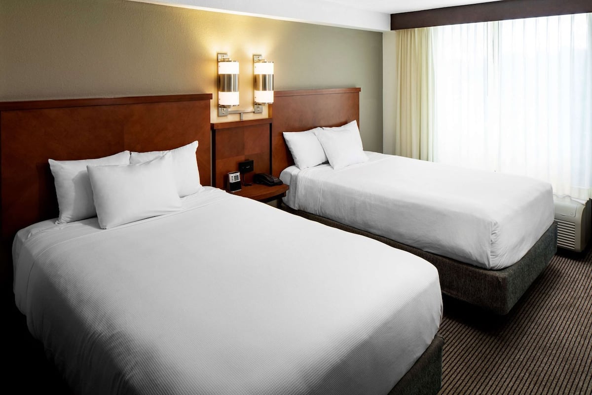A hotel room with two beds, a wooden headboard, a nightstand with a phone, and wall-mounted lights.