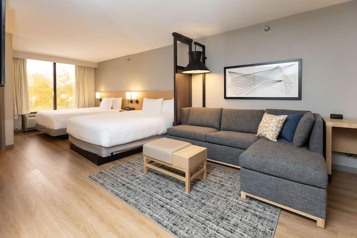 A modern hotel room with two beds, a grey sofa, a small wooden table, and a large window.