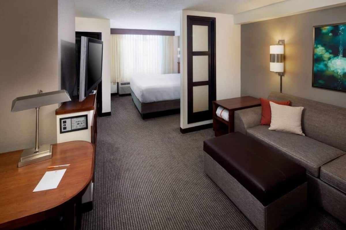 A hotel room with a bed, a desk with a lamp, a sofa, a TV, and a painting on the wall.