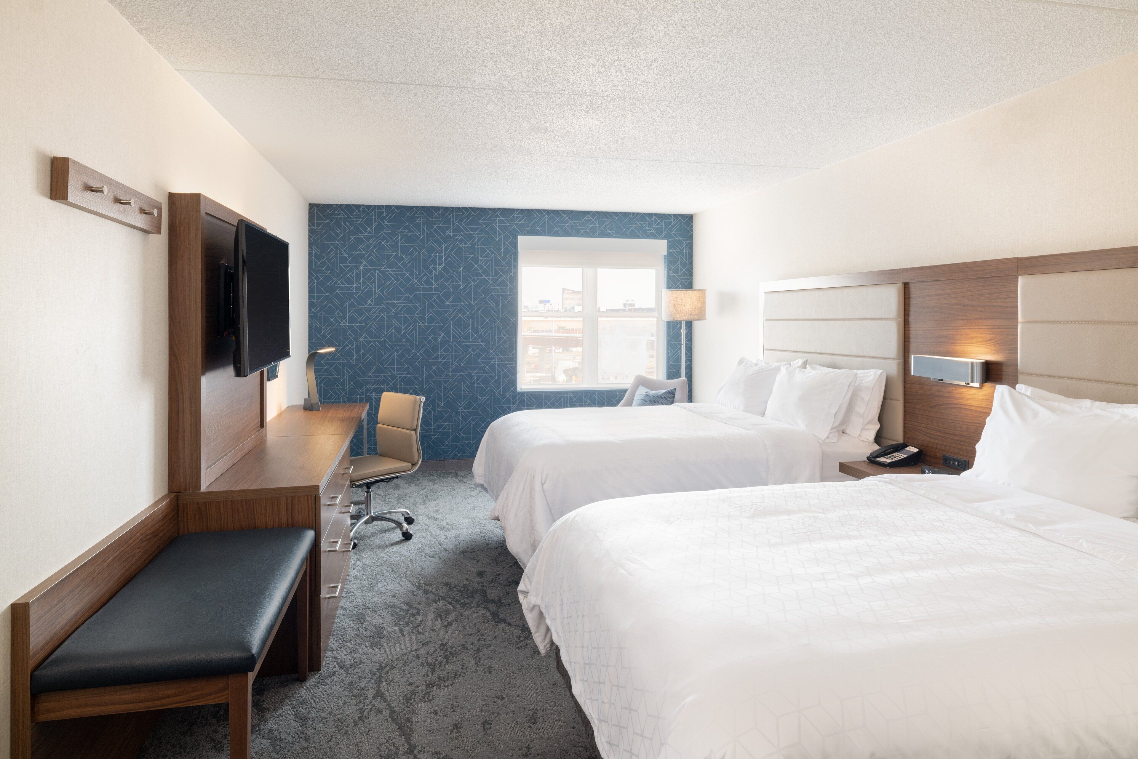 A hotel room with two beds, a desk, a bench, a TV, and a window with a city view.