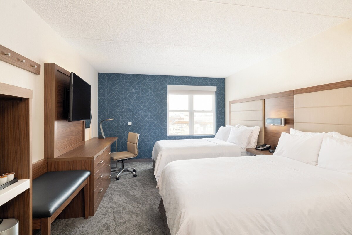 A hotel room with two beds, a desk, a chair, a TV, and a window with a view.