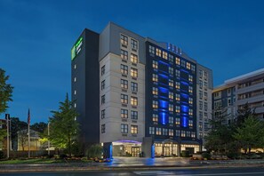 A multi-story hotel with a modern design, featuring a prominent entrance with a blue-lit canopy and a visible American flag. - Holiday Inn Express & Suites Boston - Cambridge by IHG (Cambridge)