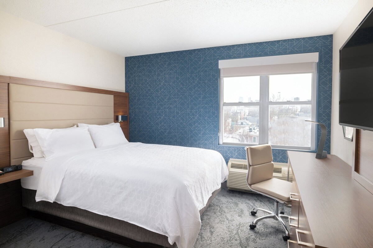 A hotel room with a bed, a desk, a chair, and a large window.
