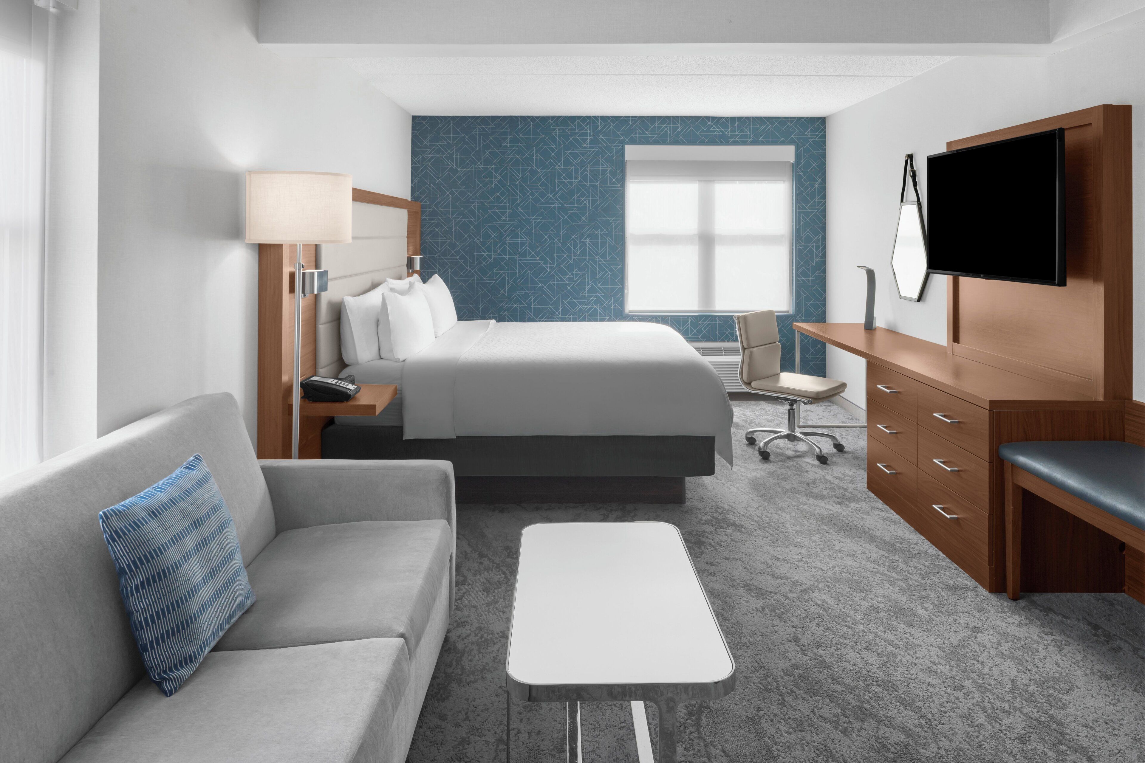 A modern hotel room with a bed, a sofa, a desk, a TV, and a large window.