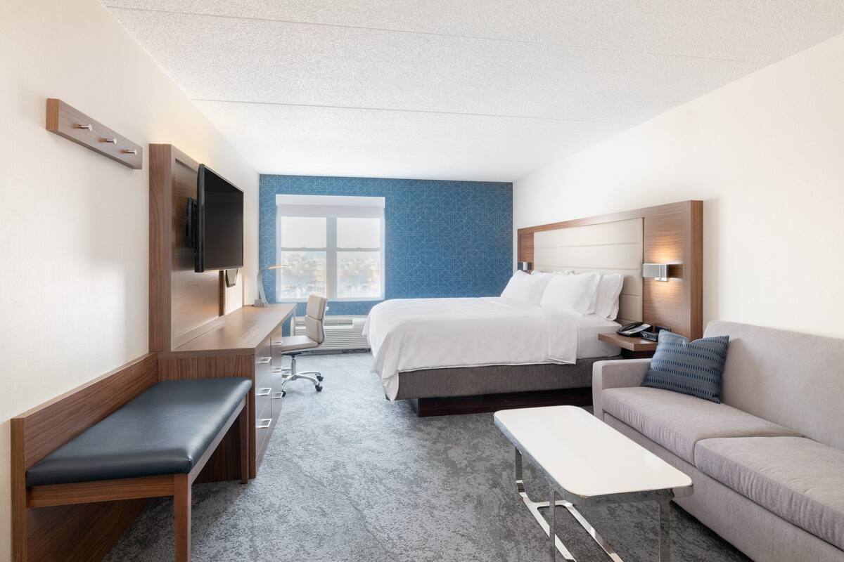 A hotel room with a bed, a sofa, a desk, a chair, a TV, and a window with a view.