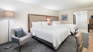 A hotel room with two beds, a sitting area with a chair, a lamp, and a framed picture on the wall.