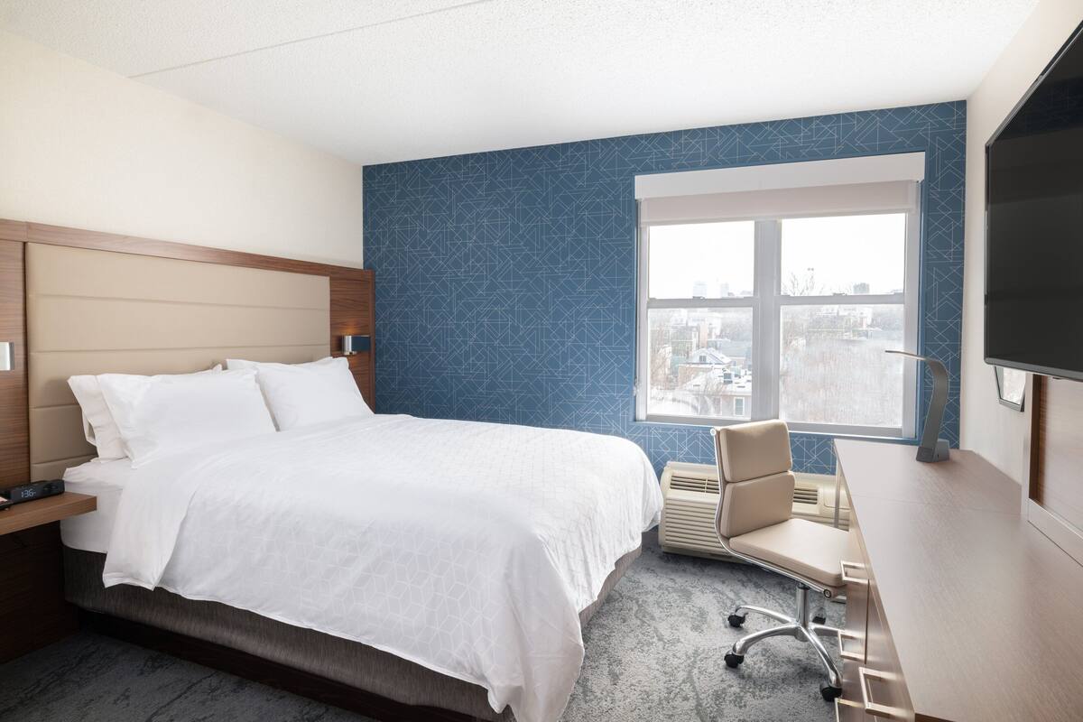 A hotel room with a bed, a desk, a chair, and a large window.