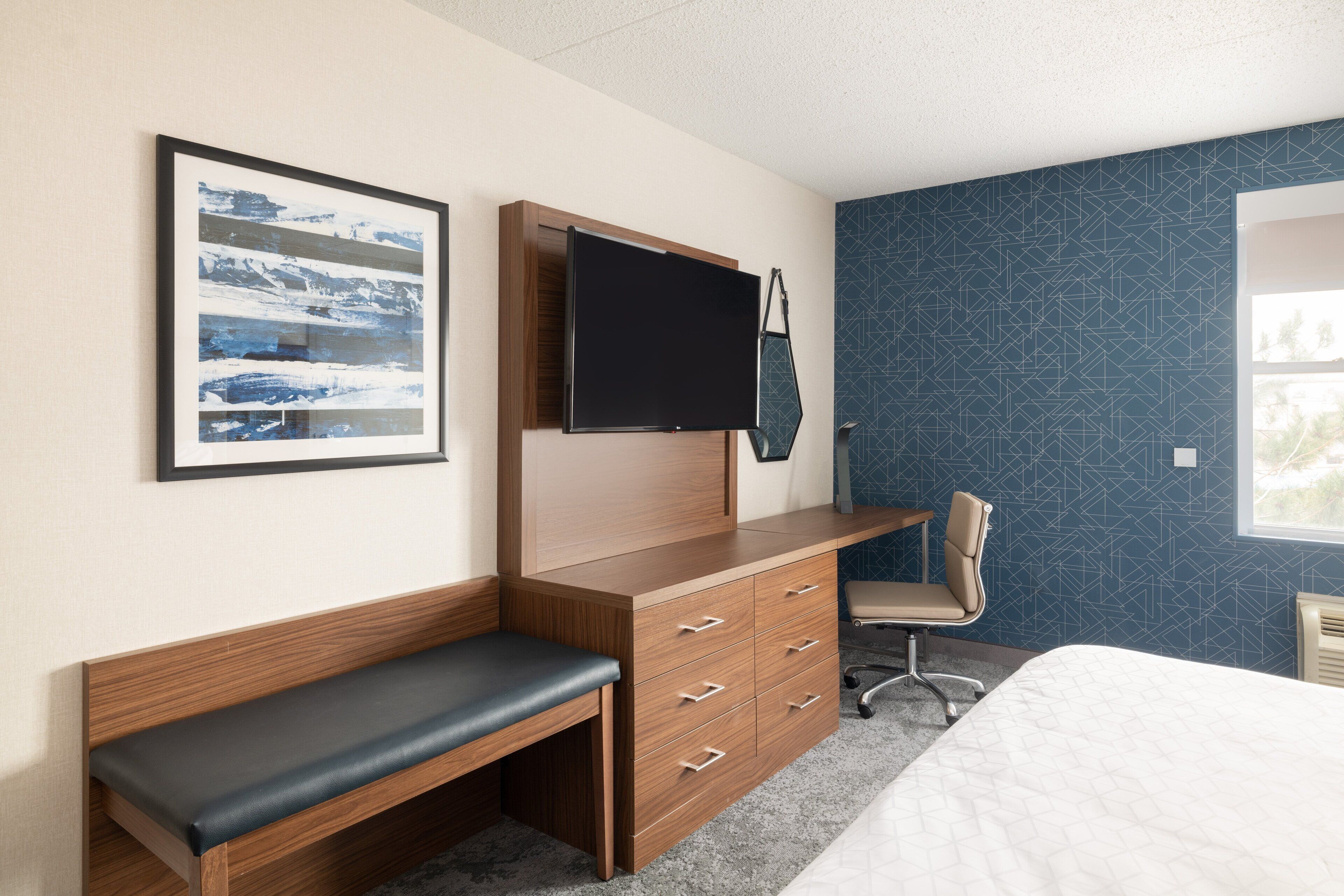 Una habitación de hotel con un escritorio de madera, un banco, un televisor montado en la pared y un papel tapiz estampado.