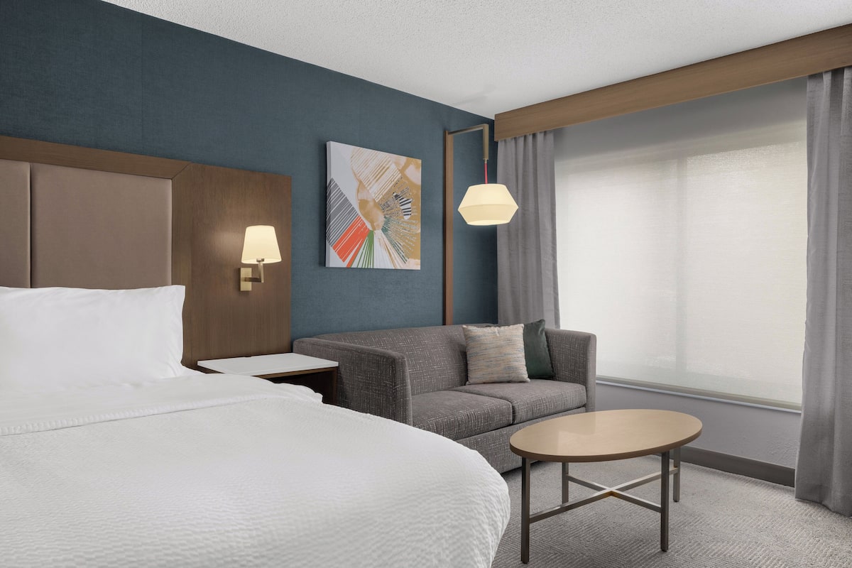 A hotel room with a bed, a sofa, a small table, a lamp, a painting on the wall, and a window with curtains.