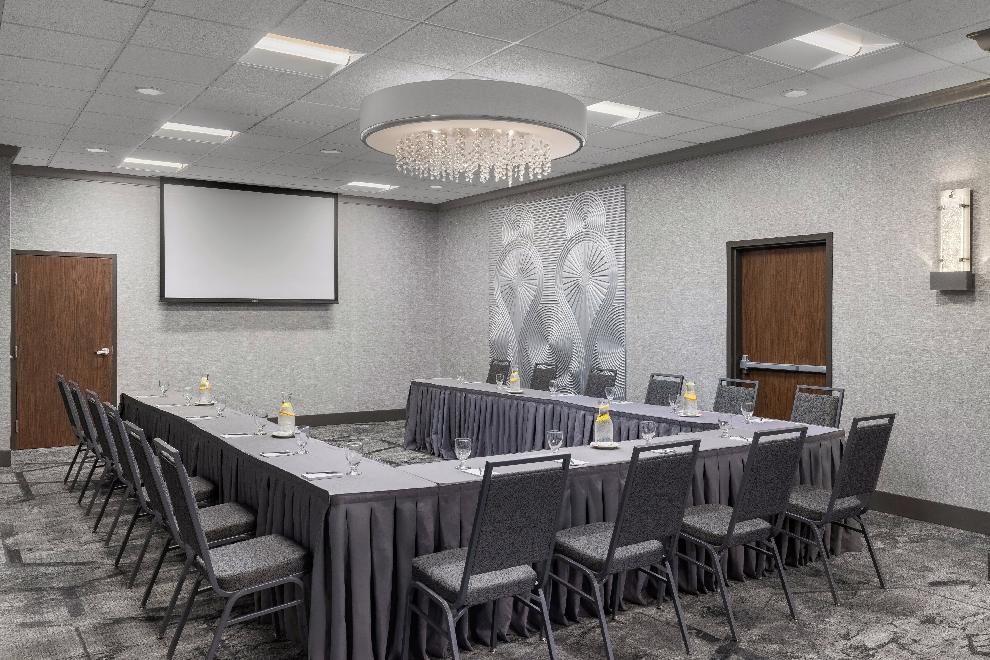 A conference room with a long table, chairs, a projector screen, and a decorative wall panel.