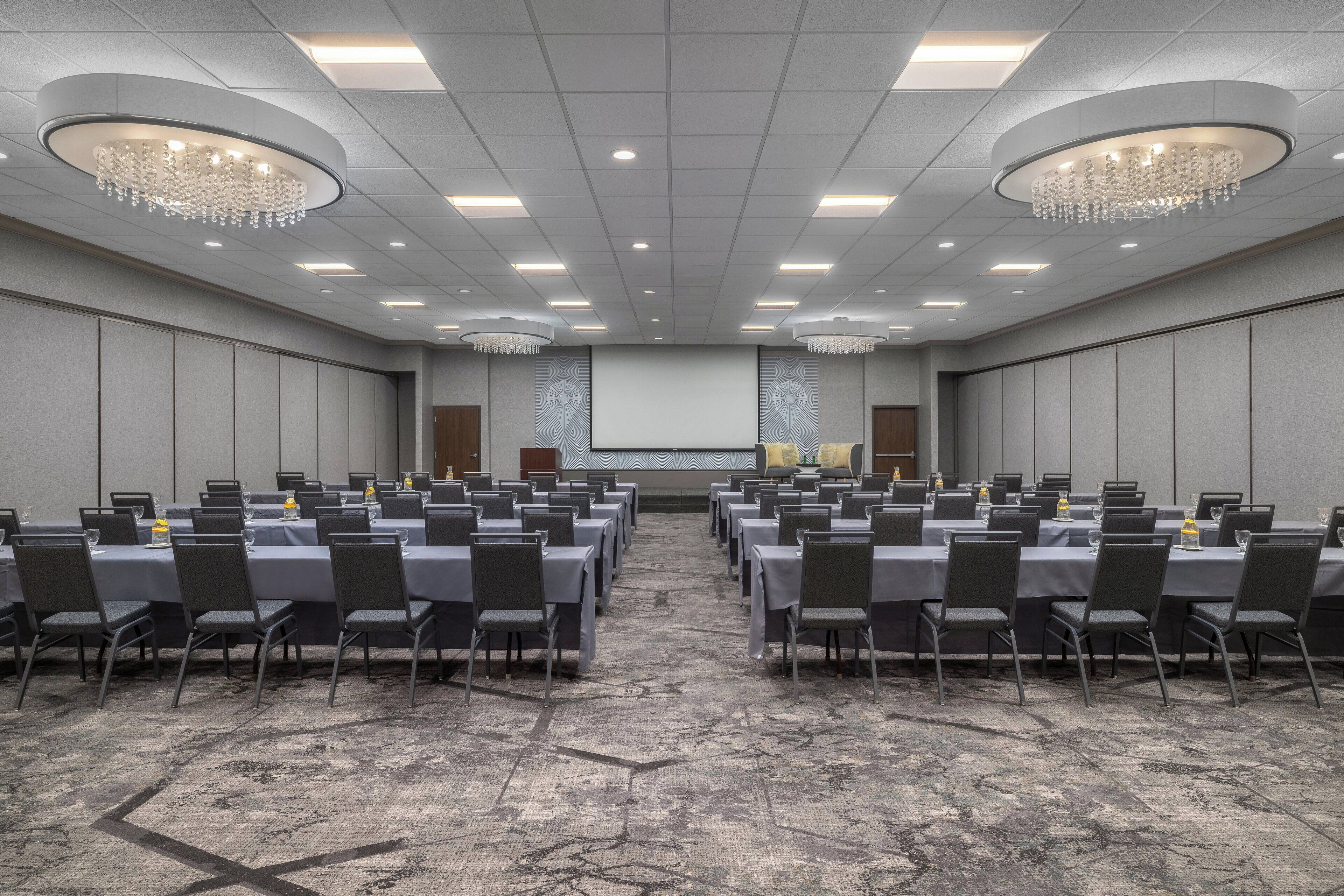 A conference room with rows of tables and chairs, a large screen at the front, and chandeliers on the ceiling.