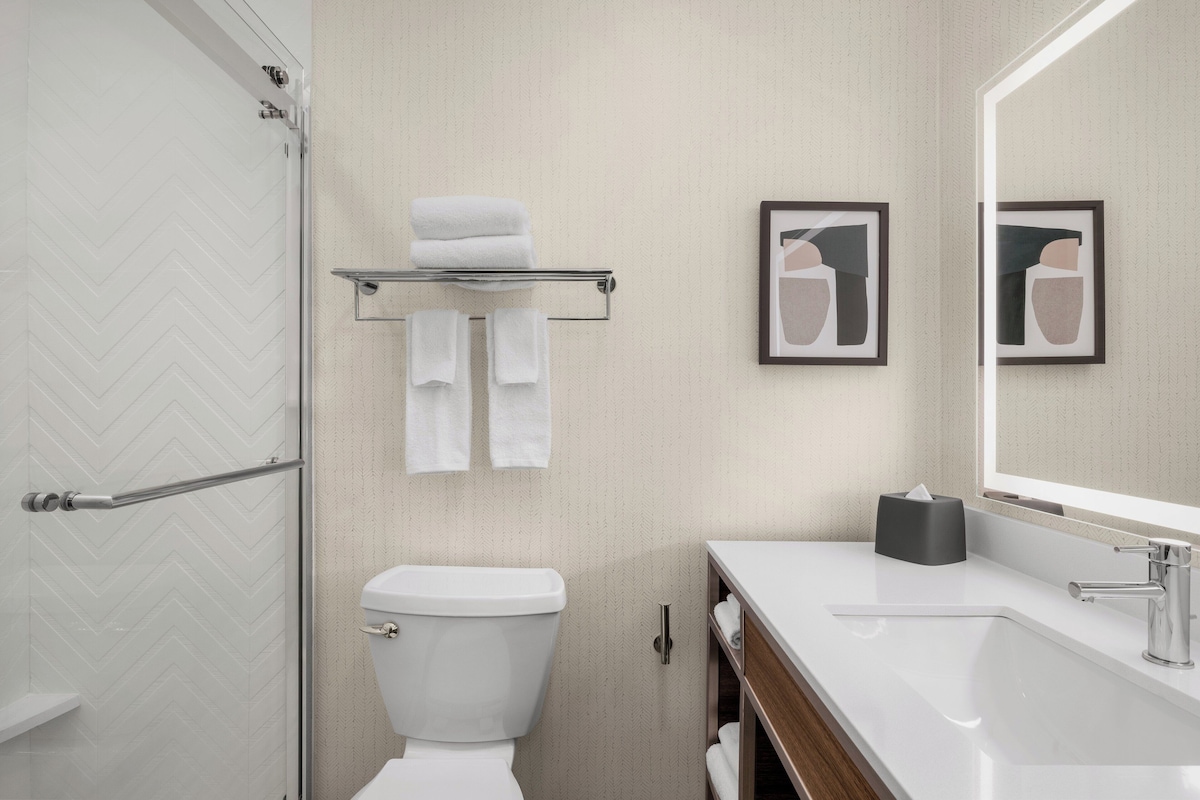 Un baño moderno con inodoro blanco, cabina de ducha de vidrio, lavabo blanco con espejo y dos cuadros en la pared.