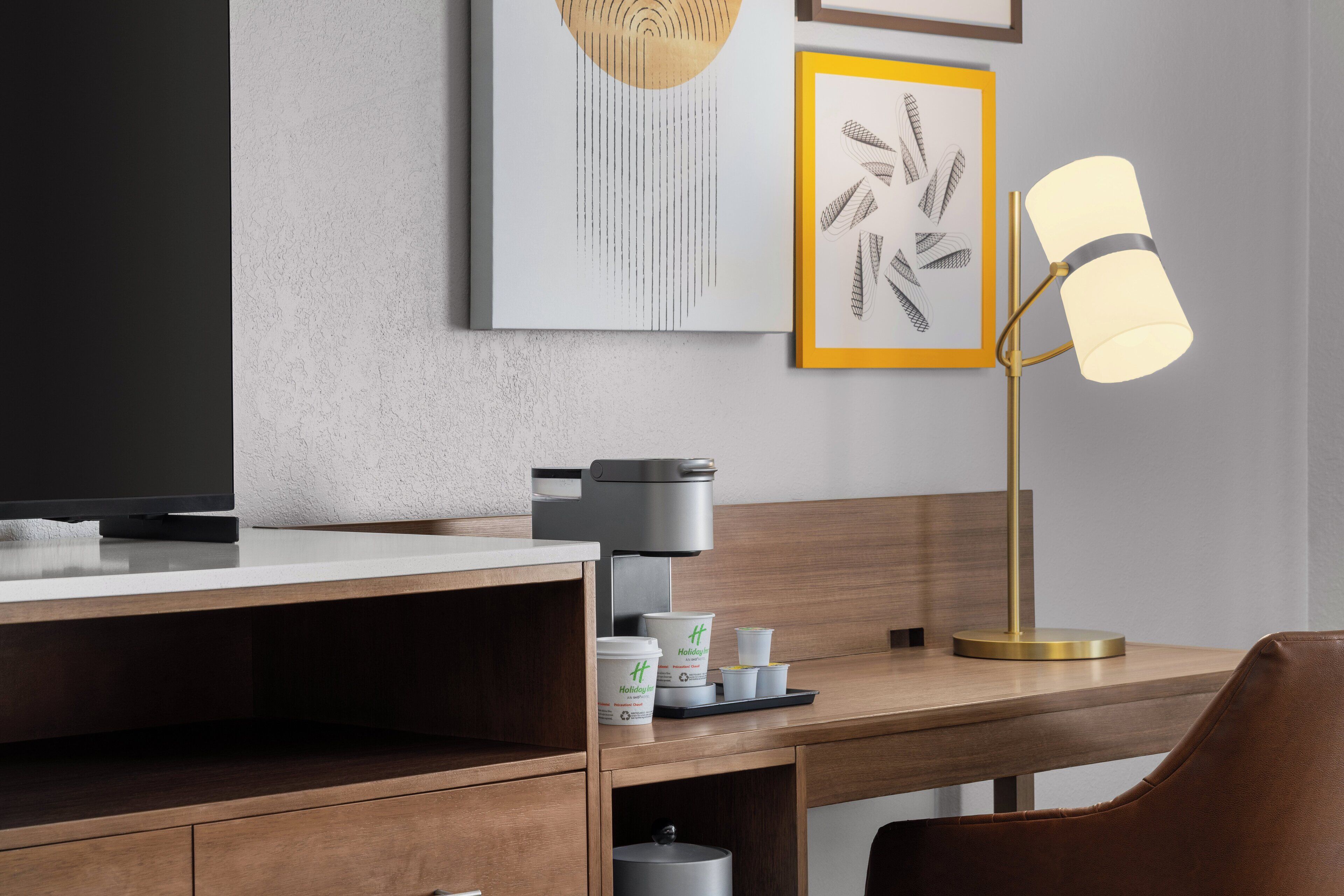 Una habitación de hotel moderna con un escritorio de madera, una cafetera, una lámpara y obras de arte enmarcadas en la pared.