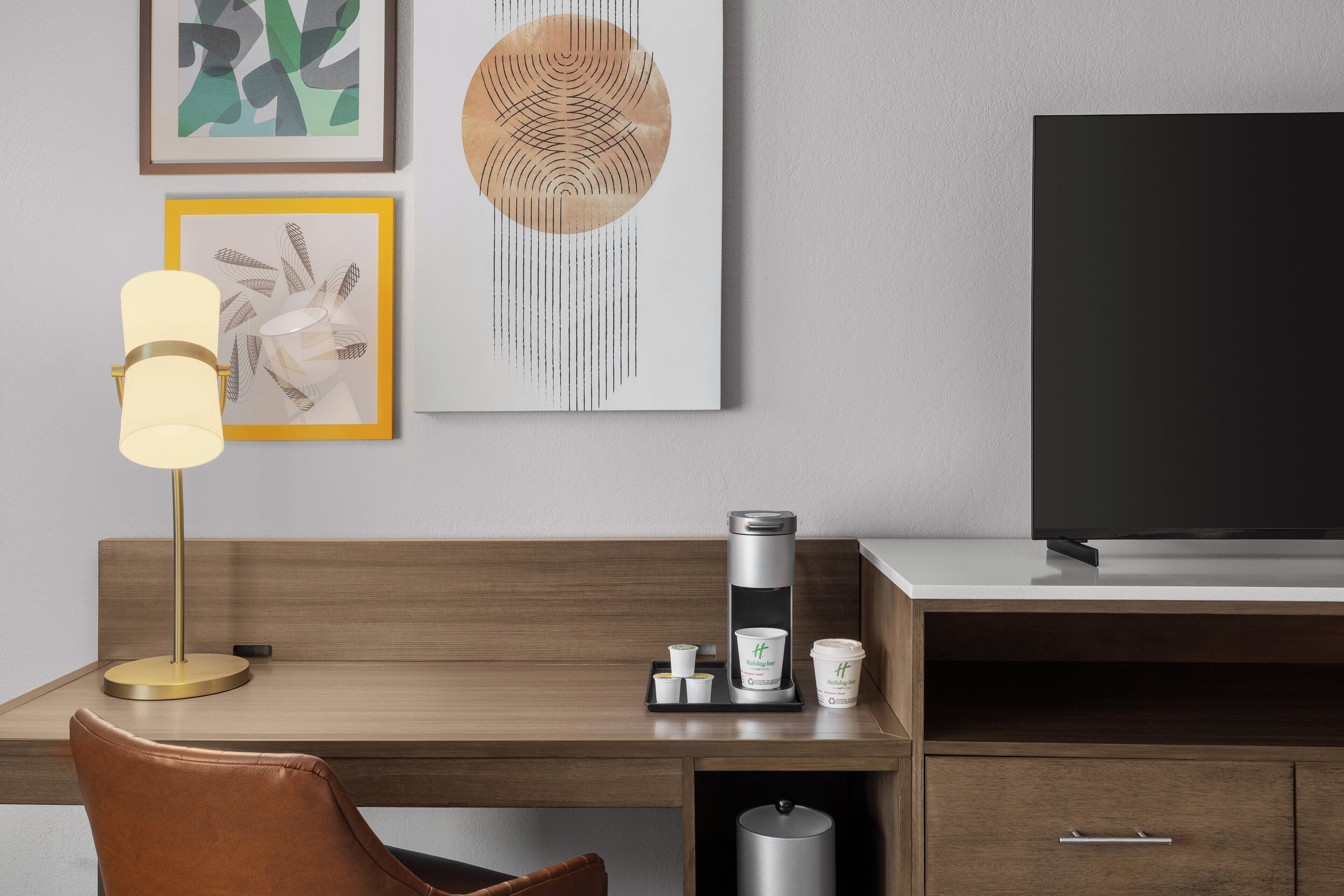 A modern hotel room with a wooden desk, a leather chair, a coffee maker, and a TV on a stand.