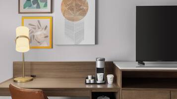 A modern hotel room with a wooden desk, a leather chair, a coffee maker, and a TV on a stand.