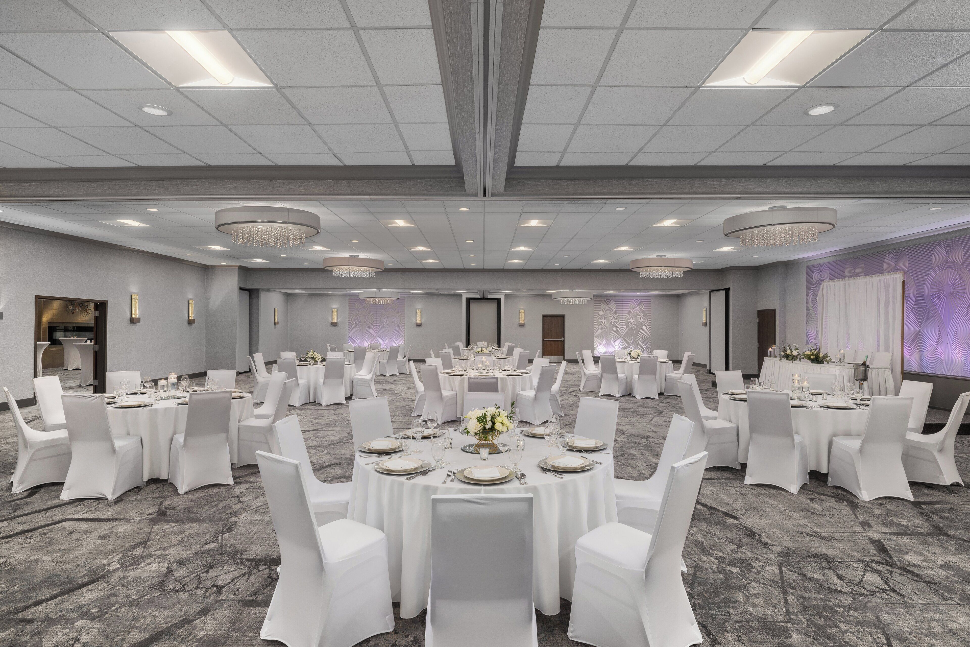 Un amplio y luminoso salón de banquetes con varias mesas redondas dispuestas para cenar, cada una rodeada de sillas blancas.