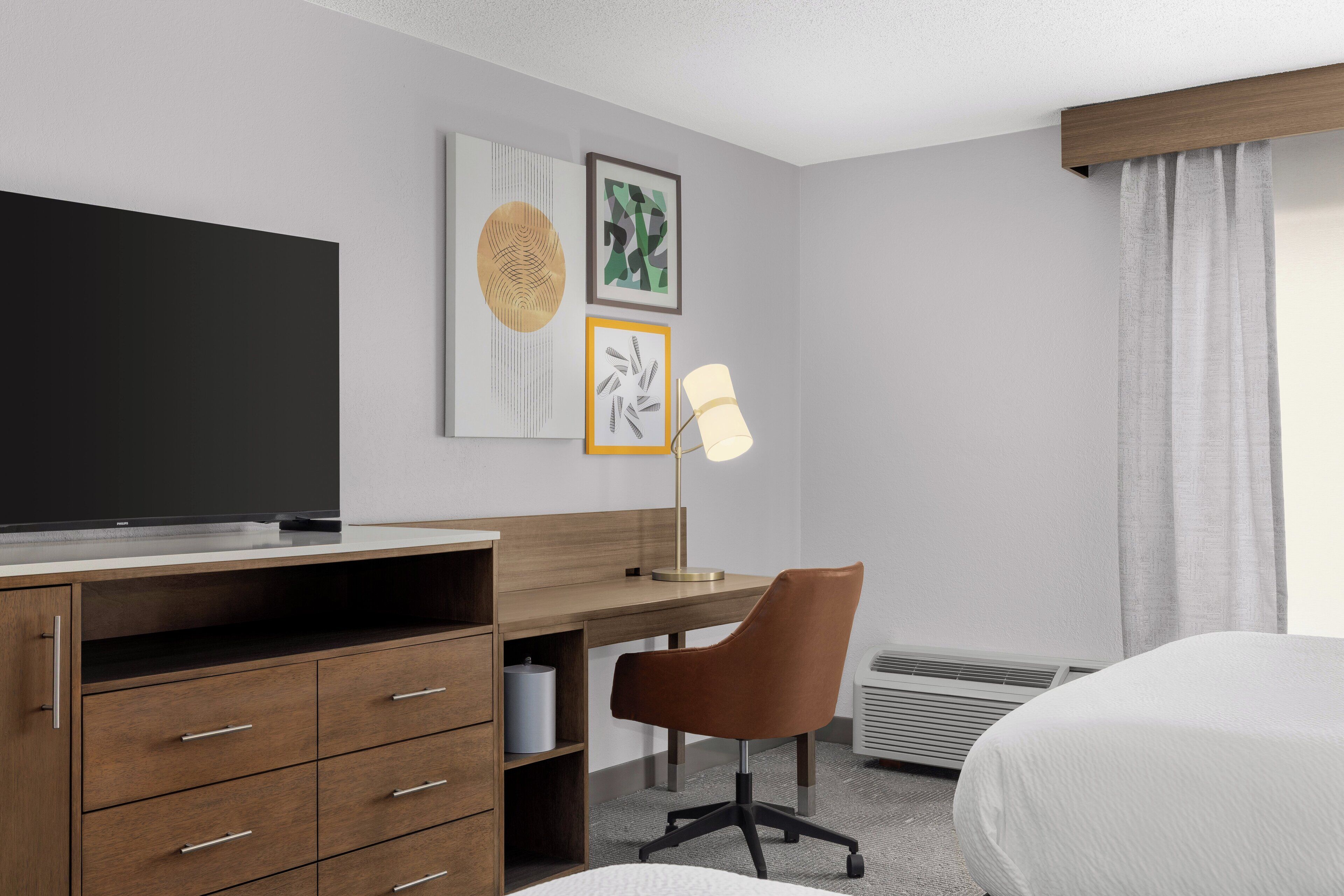 Habitación de hotel con escritorio de madera, una silla marrón, una cama, televisor y cuadros en la pared.