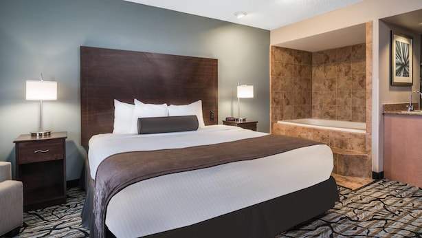 A hotel room with a large bed, two bedside lamps, a nightstand, a bathroom with a bathtub, and a patterned carpet. - Days Inn & Suites by Wyndham Wausau (Wausau)