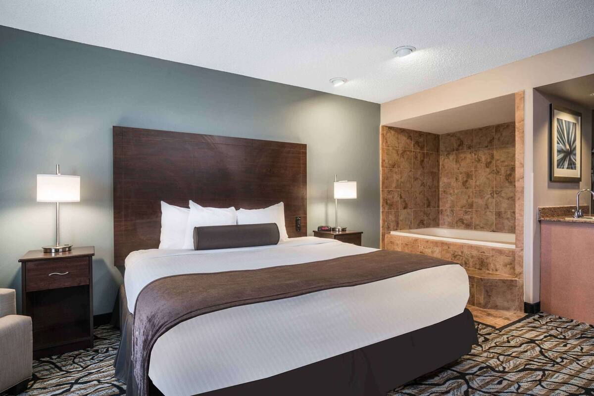 A hotel room with a large bed, bedside lamps, a bathroom with a bathtub, and a patterned carpet.