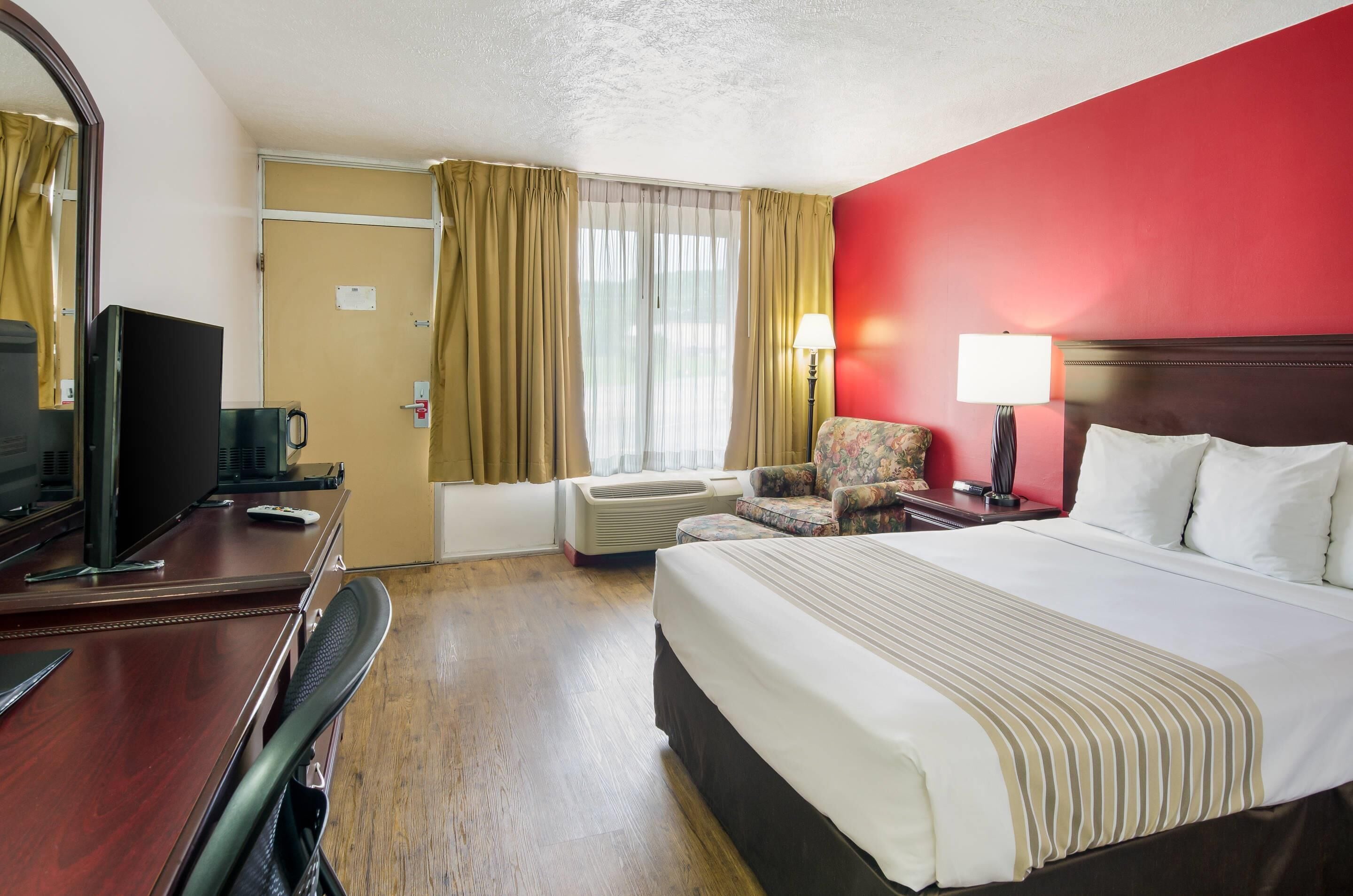 A hotel room with a bed, a desk with a computer, a chair, a TV, and a window with curtains.