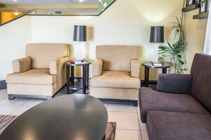 Un vestíbulo de hotel con dos sillones color beige, un sofá marrón oscuro y una mesa redonda. Hay dos mesitas auxiliares con lámparas y libros. Se ve una planta en maceta al fondo.