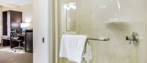 Un baño de hotel con puerta de ducha de vidrio, un toallero con una toalla blanca, un escritorio con lámpara y una silla.