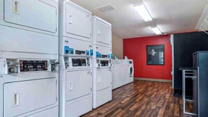 A laundry facility with multiple washing machines and dryers, a red wall, and a clock. - Studio 6 Extended Stay Huntsville, AL - US Space & Rocket Center (Huntsville)