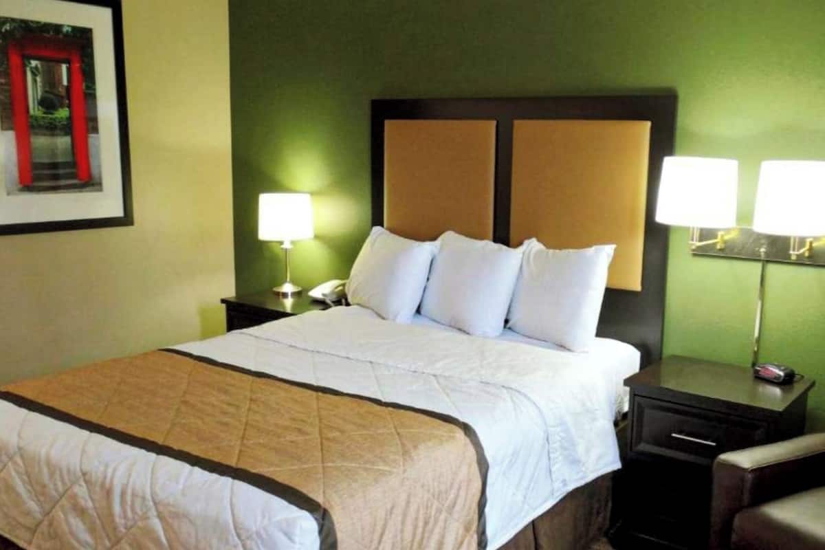 Habitación de hotel con cama, dos mesitas de noche, una silla, una lámpara y un cuadro en la pared.