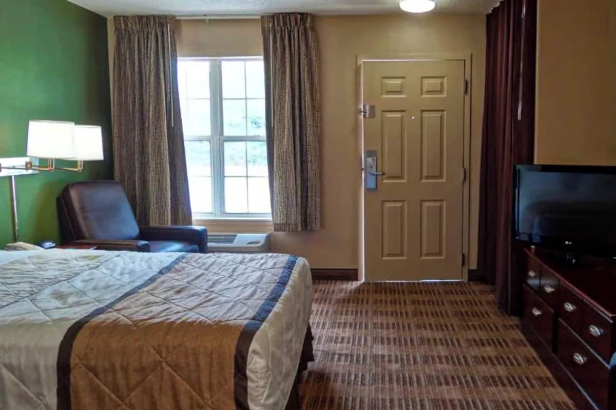 Una habitación de hotel con cama, televisor, escritorio con lámpara y ventana con cortinas.