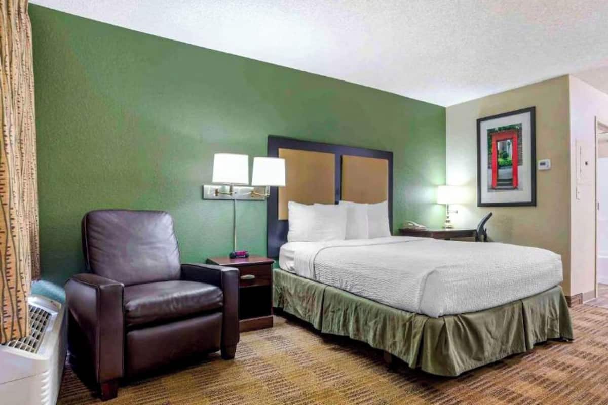 Una habitación de hotel con cama, una silla, un escritorio con lámpara y un cuadro en la pared.