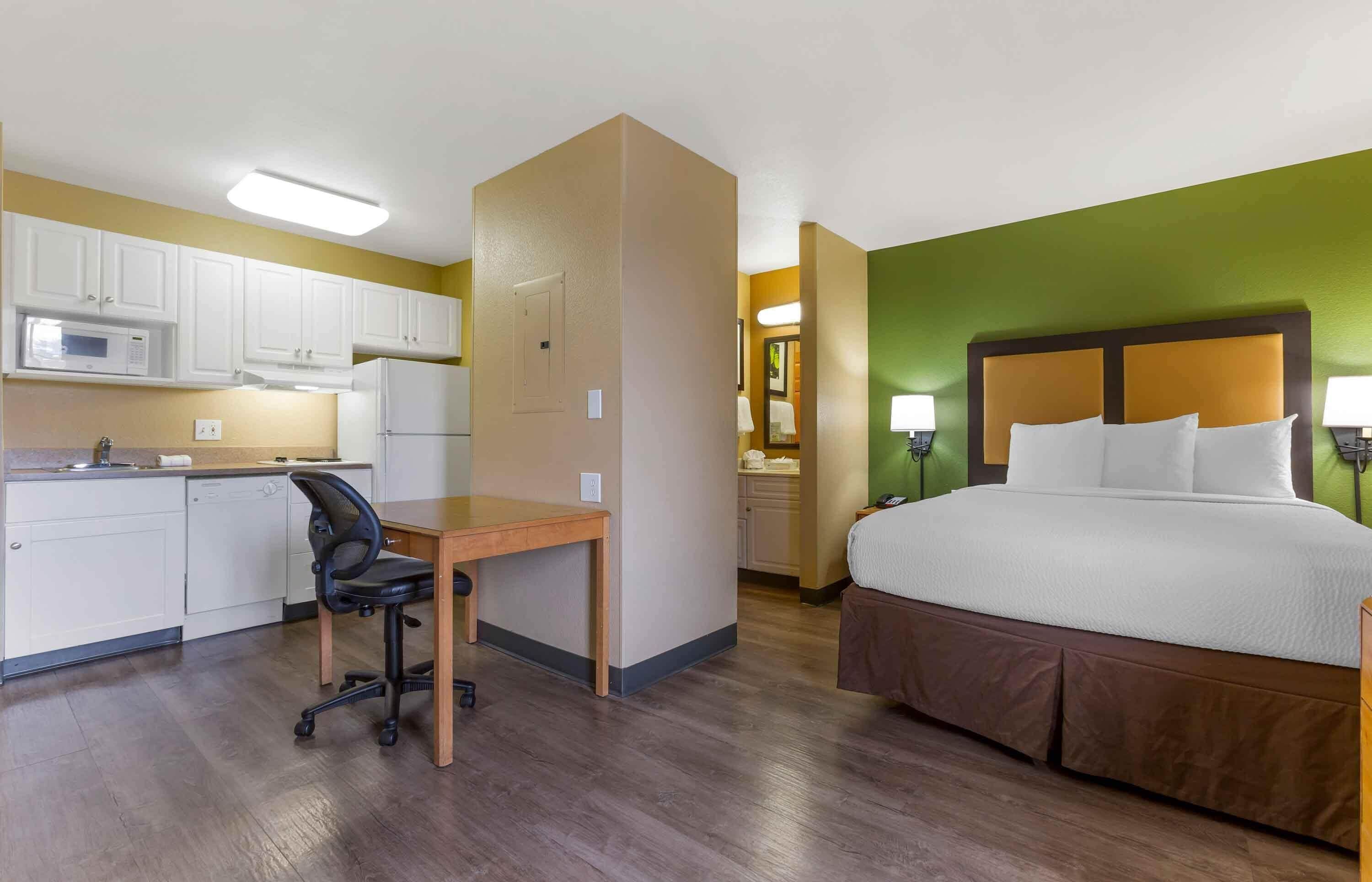 A hotel room with a bed, a desk with a chair, a kitchenette, and a bathroom.