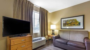 A hotel room with a flat-screen TV on a wooden cabinet, a sofa, a table with a lamp, and a framed picture on the wall.