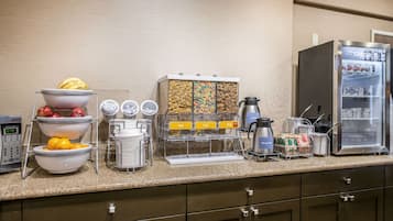 Uma mesa de pequeno-almoço com tigelas de fruta, uma máquina de café e um frigorífico.