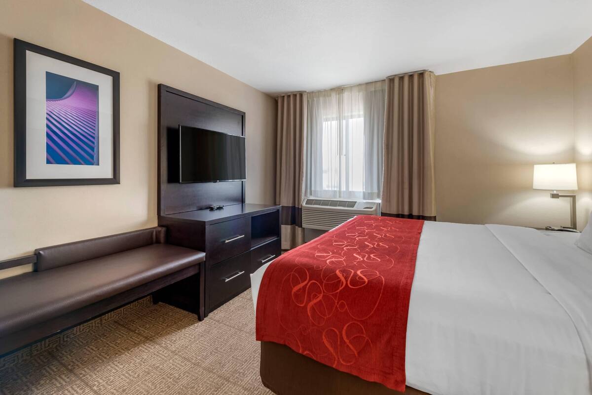 A hotel room with a bed, a desk with drawers, a flat-screen TV mounted on the wall, a bench, a window with curtains, and a wall-mounted air conditioning unit.