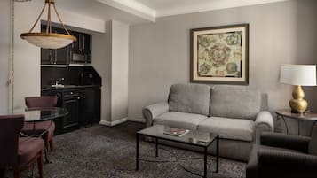 Una sala de estar con un sofá gris, una mesa de centro de vidrio y un comedor con una mesa redonda y sillas. Hay una cocina con armarios negros y un microondas. En la pared hay un cuadro con un mapa, y una lámpara sobre una mesita auxiliar.